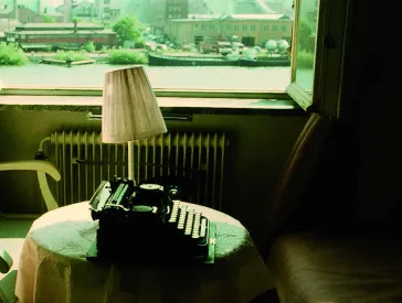 Vor dem geöffneten Fenster sieht man eine Schreibmaschine auf einem kleinen Tisch und eine Lampe mit schiefsitzendem Schirm