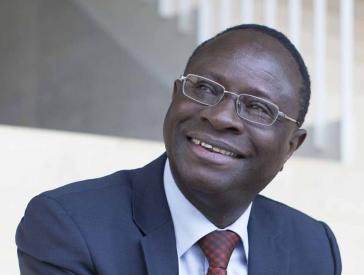 Karamba Diaby is sitting on a staircase, wearing a blue suit with a red check tie.