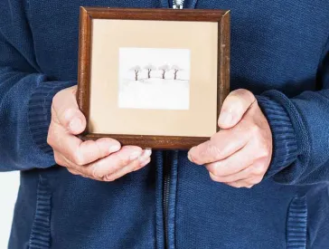 Älterer Herr hält ein gezeichnetes Bild mit vier Bäumen in einem Rahmen in Händen.