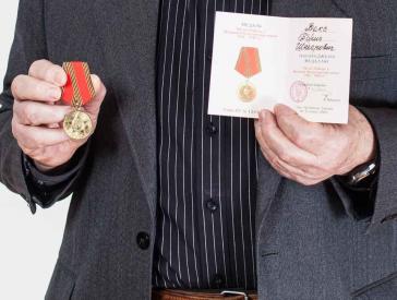Older gentleman in dark jacket holds medal and a document in his hands.