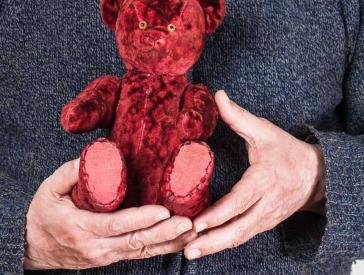 Elderly man holding a red teddy bear in his hands.