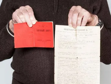 Ein bärtiger grauhaaruger Mann hält zwei Dokumente hoch.