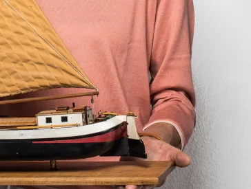 Hände halten ein Holzbrett, auf dem ein Modellschiff steht.