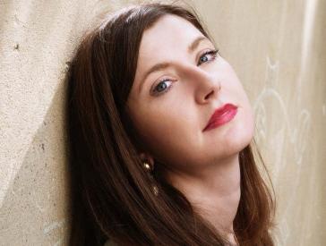 Woman with long brown hair and red lipstick leans her head against a wall and looks at the camera