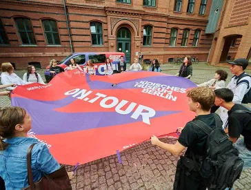 Eine Gruppe von Jugendlichen spannen auf einem Innenhof ein großes Tuch auf, auf dem on.tour und Jüdisches Museum Berlin steht.