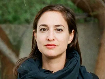 A woman with long brown hair stands in front of a green background and looks into the camera.