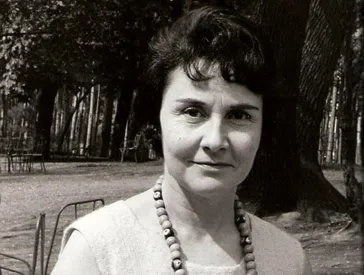 Schwarz-Weiß-Foto einer mittelalten Frau mit kurzen dunklen Haaren und auffälliger Kette um den Hals, die freundlich-skeptisch in die Kamera schaut, im Hintergrund Bäume.