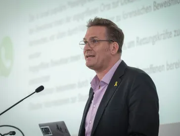 Ein Mann mit kurzen Haaren, Brille und gelber Schleife am Revers seines Sakkos steht vor einer Leinwand und spricht in ein Mikrofon.