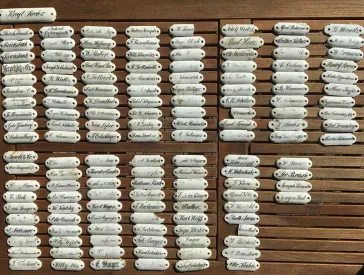 Auf einem Holztisch sind zahlreiche kleine weiße Namensschilder aus Porzellan aufgereiht. 