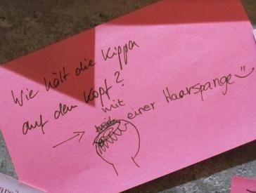 Post-it note in pink: "Wie hält die Kippa auf dem Kopf? - mit einer Haarspange" (How does a kippah stay on? - with a hair clip))