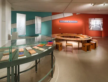Blick in einen Ausstellungsraum, im Vordergrund eine runde grüne Vitrine mit Büchern, im Hintergrund ein runder Holztisch mit Stühlen, dahinter eine orange-gestrichene Wand mit einem Schild des Kaufhauses Schocken.