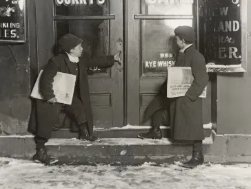 Schwarz-weiß Fotografie von zwei Kindern, die mit Zeitungen unter dem Arm vor einer Tür stehen.