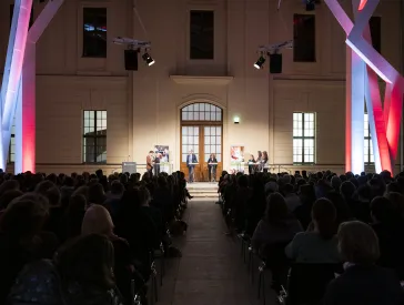 Im Glashof im Jüdischen Museum Berlin sind die Zuschauerränge gefüllt. Erhöht stehen Menschen an Pulten. Die Balken sind bunt beleuchtet.