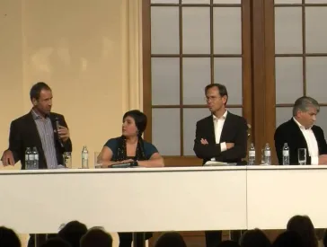 Drei Personen sitzen an einem Tisch auf einer B&uuml;hne.