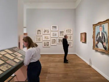 Ausstellungsraum mit Besucher*innen und Gemälden an weißer Wand, einer Vitrine mit Gemälden und Zeichnungen in der Mitte des Raumes.
