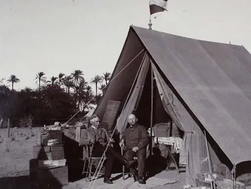 Schwarz-weiß Fotografie, zwei Männer in Anzügen sitzen vor einem Zelt.