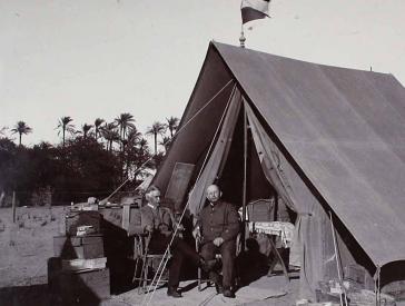 Schwarz-weiß Fotografie, zwei Männer in Anzügen sitzen vor einem Zelt.
