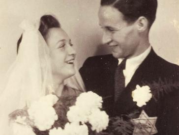 Black and white photograph of a couple with a bouquet of flowers and a veil, a yellow badge on his jacket.