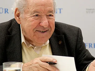 Photography: a smiling older man holds a pen in his hand and sits in front of an open book.