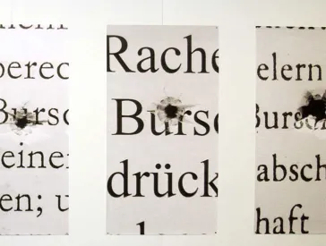 An einer Wand hängende Plakate, auf denen groß aufgezogene Wörter und Einschusslöcher zu sehen sind