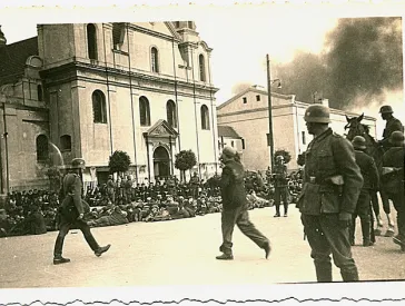 Schwarz-wei&szlig; Fotografie von einer Kirchenfassade vor der Menschen auf dem Boden sitzen, im Vordergrund deutsche Soldaten.