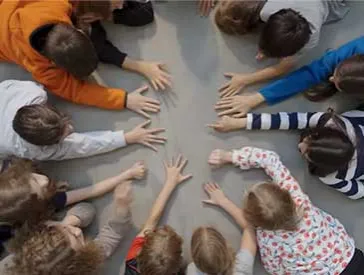 Photo of about ten children. We see them from above, lying face down on the floor and forming a circle. Their heads are facing the center of the circle, and most of them are stretching one arm toward the center.