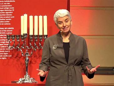 A woman with short gray hair (Hetty Berg) stands in front of a Hanukkah menorah.