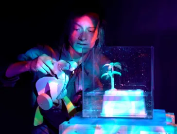 An actress holds a wooden toy in her hands and stands next to a white glass box containing a plastic palm tree. It is dark, the actress and the objects are illuminated by coloured light.