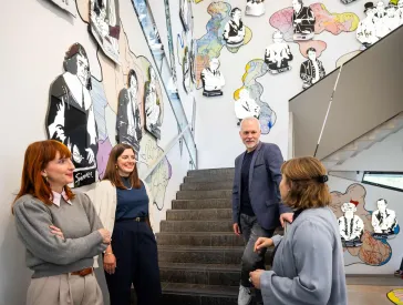 Vier Menschen stehen einander zugewandt an einer Treppe, im Hintergrund an den W&auml;nden Comic-artig stilisierte ber&uuml;hmte j&uuml;dische Pers&ouml;nlichkeiten.
