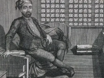 A man with a blank stare sits on a bed in a cell.