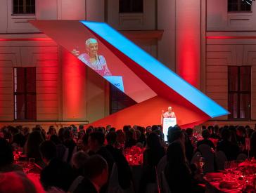 A red-lit stage, with a woman in a pink dress in front of a lectern.