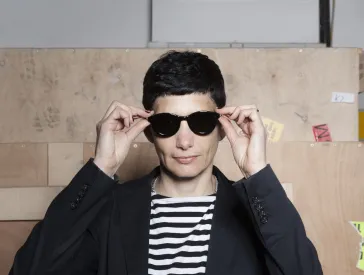 Portrait (bust) of a woman with black short hair. She wears a black jacket and a black and white cross-striped T-shirt and is putting on dark sunglasses.