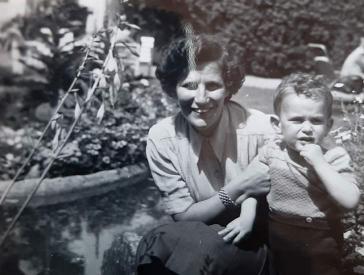 Schwarz-weiß-Foto eines Kleinkinds in Lederhosen, daneben kniet eine Frau in Rock und Bluse, im Hintergrund ein Haus und viele Pflanzen.