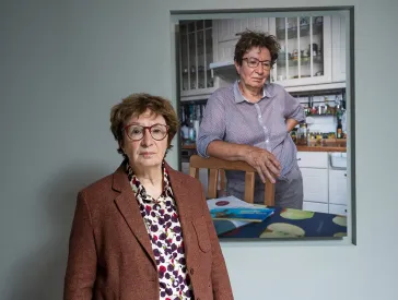 Eine Frau in Bluse und braunem Jackett steht in der Ausstellung vor ihrem Porträt, das in einer Küche aufgenommen ist