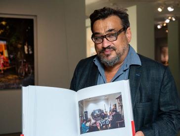Ein Mann mit Bart und Brille steht in einer Fotoausstellung und hält einen aufgeschlagenen Katalog mit einem Bild von sich in der Hand