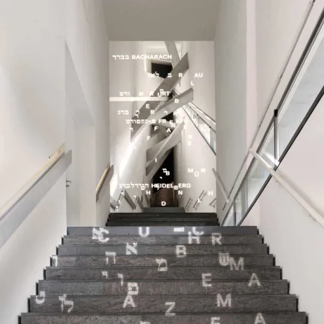 Eine schwarze hochführende Treppe, auf der weiße lateinische und hebräische Buchstaben projiziert sind.