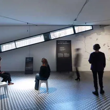 Das Bild zeigt einen Ausstellungsraum, in dem weiße Hocker verteilt sind, auf denen Menschen sitzen. Andere Menschen bewegen sich durch den Raum. Es gibt Aufsteller und Wände mit Text und einer Grafik..