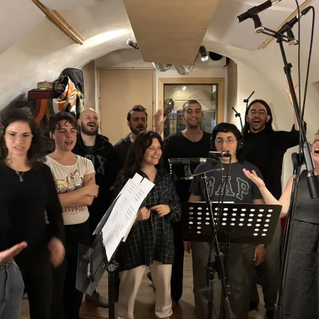 Eine Gruppe von Leuten steht zusammen in einem Raum. Sie singen zusammen. Vor ihnen stehen Notenst&auml;nder und Mikrofone. 