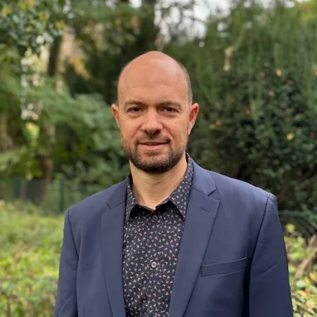 Mann schaut freundlich in die Kamera. Er tr&auml;gt ein blaues Jackett und ein gemustertes Hemd.