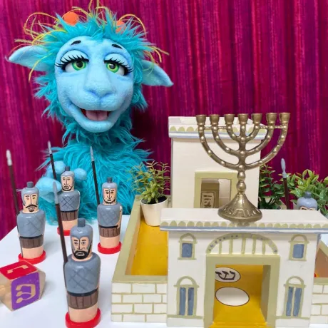 Eine blaue zottelige sprechende Puppe mit großen schönen Augen hinter einem Tisch mit Spielzeugfestung, Dreidel und Chanukka-Leuchter darauf.