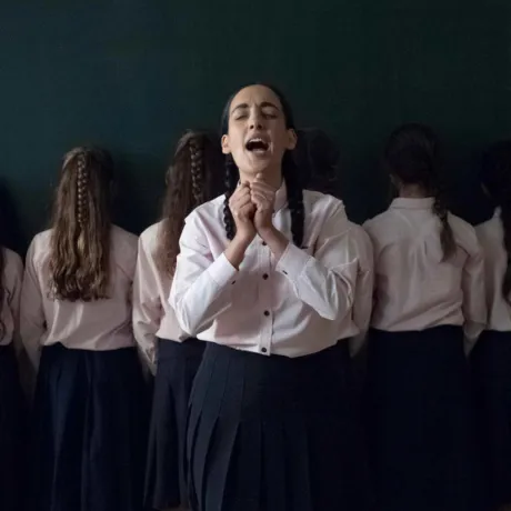 Several young women stand in a row with their backs to the viewer. One young woman stands slightly in front of the row, facing the camera. Her eyes are closed, her mouth is open, and she holds her hands vertically in front of her, loosely clenched into fists so that they touch.