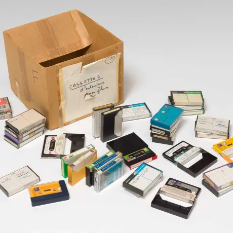 An empty, old cardboard box stands on a white background. Around the box are about 30 audio cassettes in hand-labeled cases.