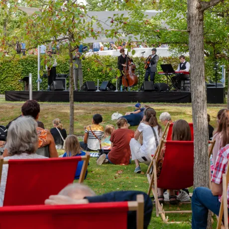 Das Publikum liegt in einem Garten auf Liegest&uuml;hlen und schaut zu einer Musikband, die auf einer B&uuml;hne spielt.