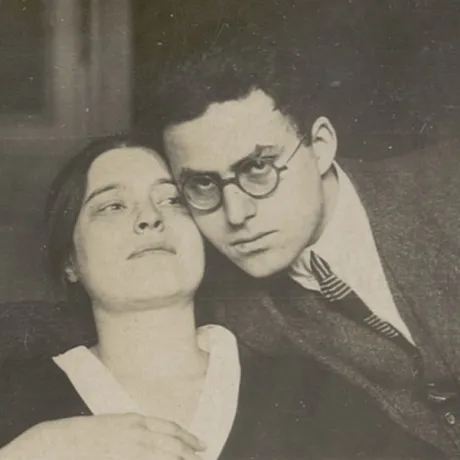 Die historische Fotografie zeigt eine junge Frau, zu deren rechter Seite ein Mann an ihren Kopf gelehnt ist. Er blickt in die Kamera und umfasst die Schultern der Frau mit seinem rechten Arm.