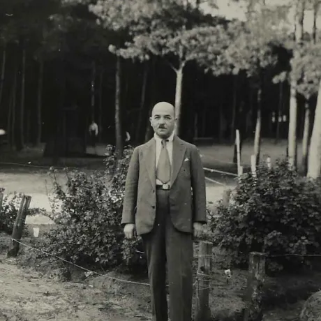 Das Bild zeigt einen Anzug tragenden Mann im Ganzk&ouml;rperportr&auml;t. Hinter ihm B&uuml;sche und B&auml;ume.