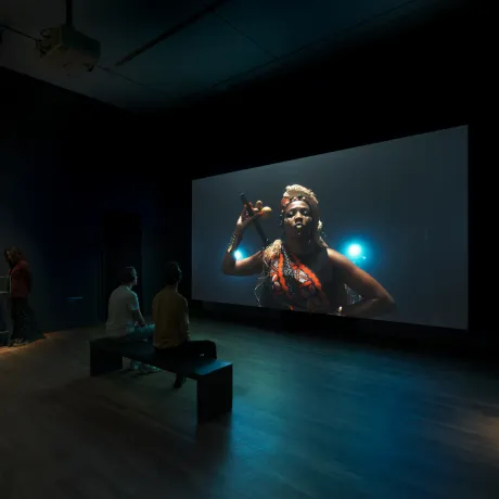 Blick in einen Ausstellungsraum mit Filmleinwand, darauf eine weiblich und karibisch gelesene Person mit rot-schwarzer Scherpe, Kopfschmuck aus Stroh und Stab mit Calebassen in der Hand vor dunklem Hintergrund.