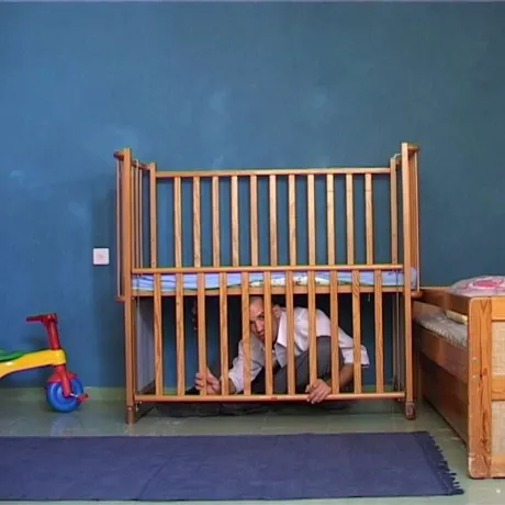 Ein Erwachsener sitzt auf dem Boden eines Kinderzimmers unter dem Gitterbett für Babys und schaut durch die heruntergelassenen Holzgitterstäbe.
