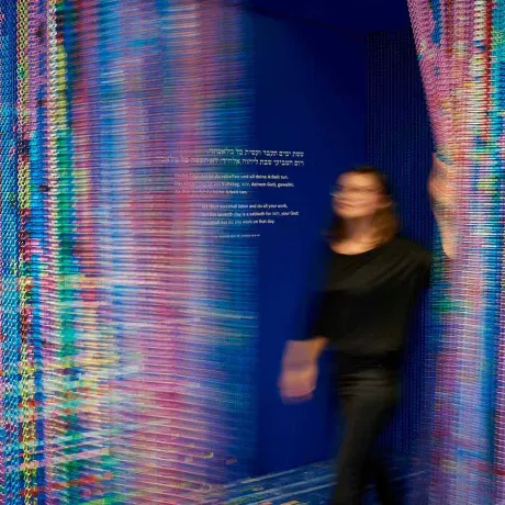 Eine Person geht durch einen bunt glitzernden Vorhang, auf der Wand das Gebot am siebten Tage zu ruhen (auf Hebräisch, Deutsch und Englisch)