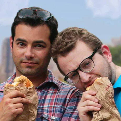 Two men in short-sleeved shirts are standing in the park, eating baked goods out of paper bags and looking at the camera.