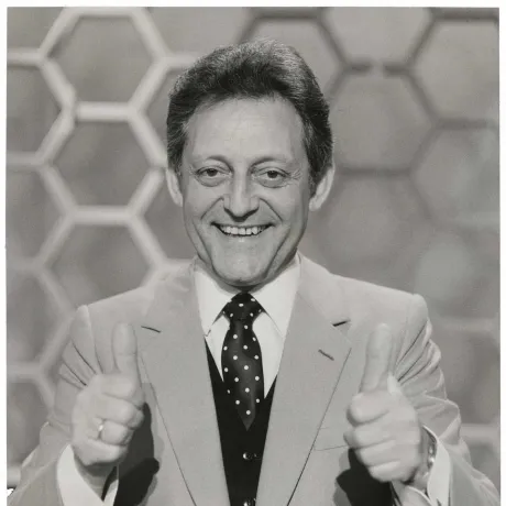 The black and white photo shows a half-portrait of a man in suit and tie with both thumbs up and looking friendly into the camera, with honeycomb pattern in the background. 